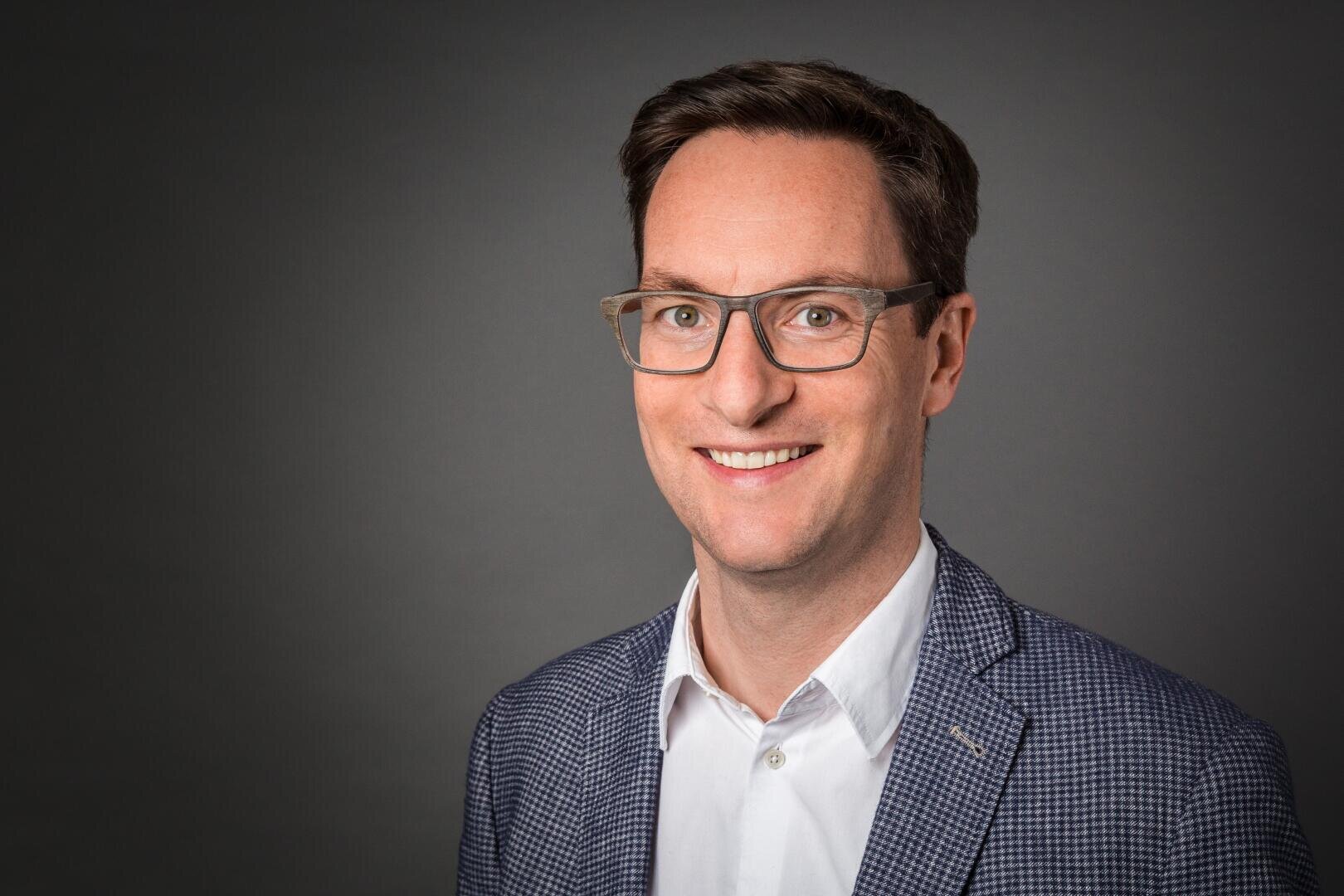 Ein Mann mit kurzen braunen Haaren und Brille, der ein weißes Hemd und einen blau gemusterten Blazer trägt, lächelt in die Kamera vor einem schlichten dunkelgrauen Hintergrund.