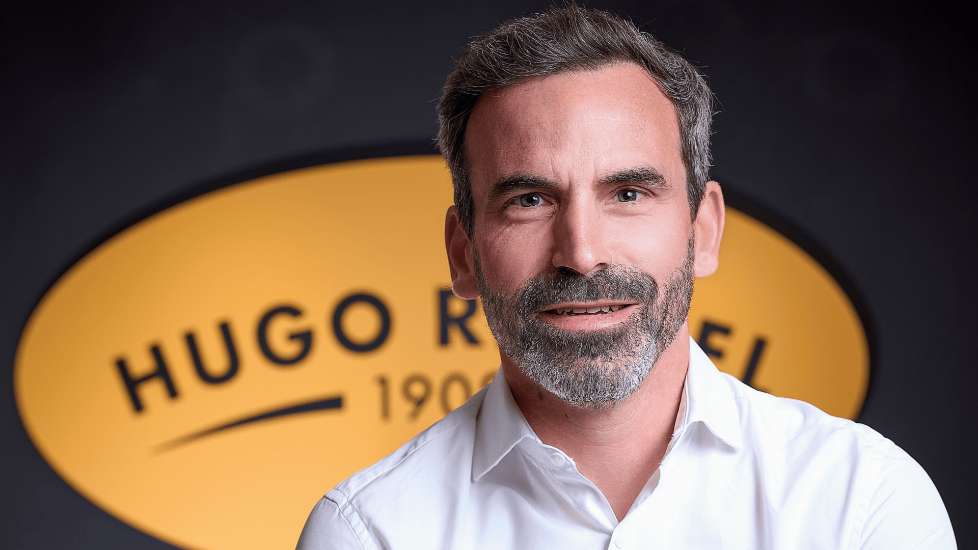 Ein Mann mit kurzen grauen Haaren und Bart, der ein weißes Hemd trägt, posiert vor einem unscharfen gelben ovalen Schild, auf dem der Text HUGO R. und 190 zu sehen ist.