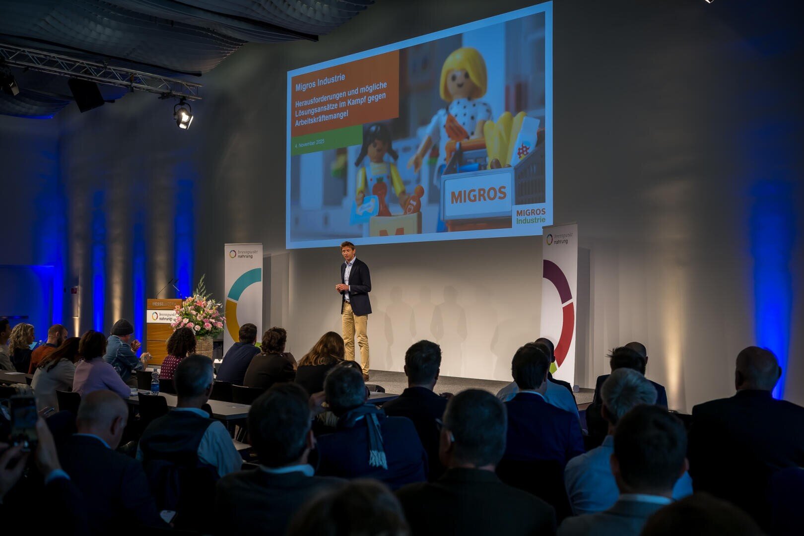 Ein Mann spricht auf der Bühne zu einem Publikum bei einer Konferenz. Auf einem großen Bildschirm hinter ihm wird eine Präsentationsfolie der Migros Industrie mit einer Playmobil-Figur, Text und dem Migros-Logo angezeigt. Der Raum ist gut beleuchtet und hat blaue Akzente.