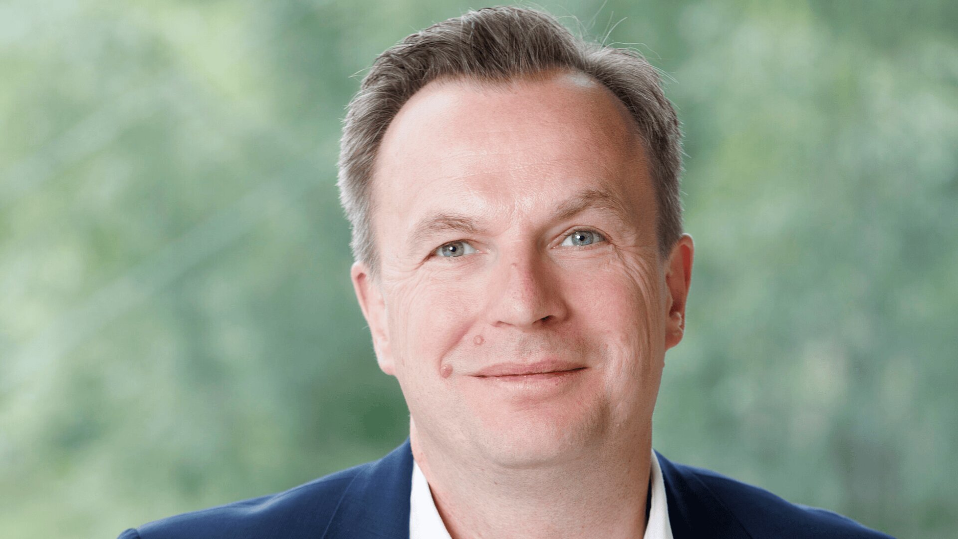 Ein Mann mittleren Alters mit kurzen braunen Haaren, heller Haut und blauen Augen lächelt sanft in die Kamera. Er trägt ein dunkles Jackett und ein weißes Hemd. Der Hintergrund ist von verschwommenem Grün geprägt.