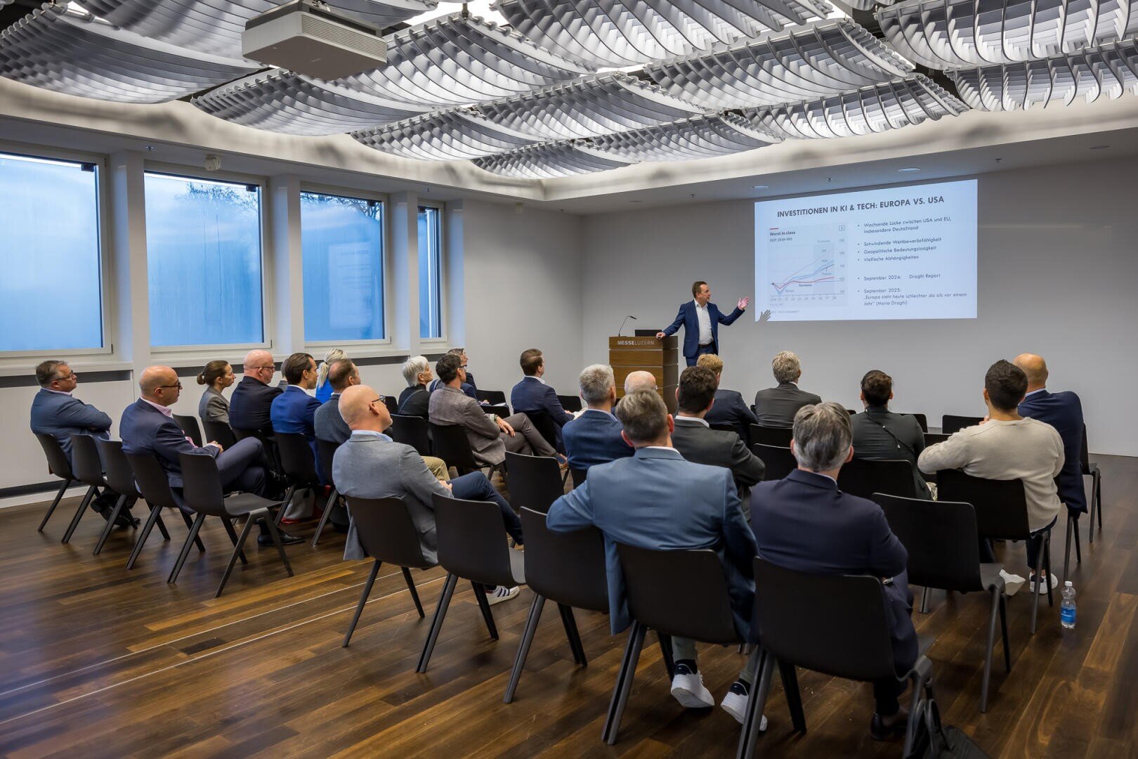 Ein Mann im Anzug hält einen Vortrag vor einer Gruppe von sitzenden Fachleuten in einem modernen Konferenzraum. Eine Folie mit dem Titel INVESTMENTS IN VC TECH: EUROPE VS USA wird an die Wand projiziert.