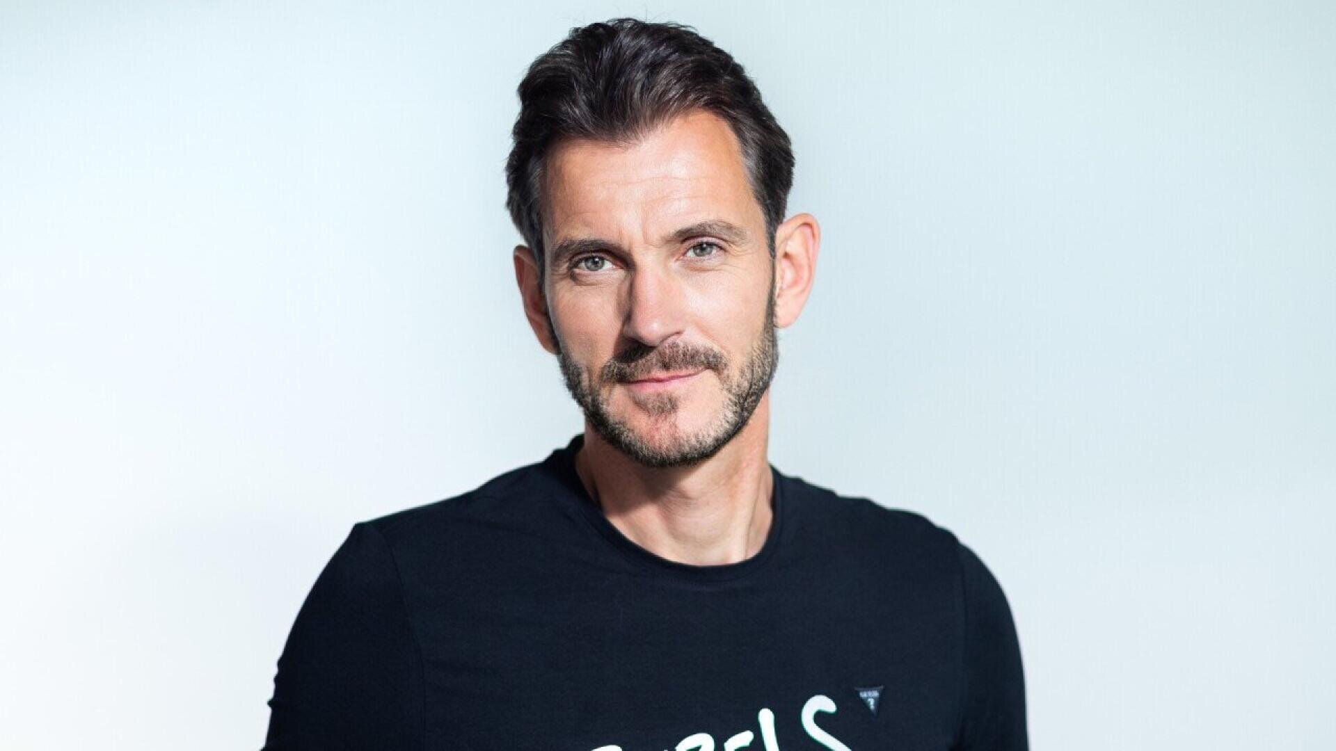 Un homme aux cheveux bruns courts et à la barbe, vêtu d'un T-shirt noir, se tient devant un fond clair et uni, regardant l'appareil photo et souriant légèrement.