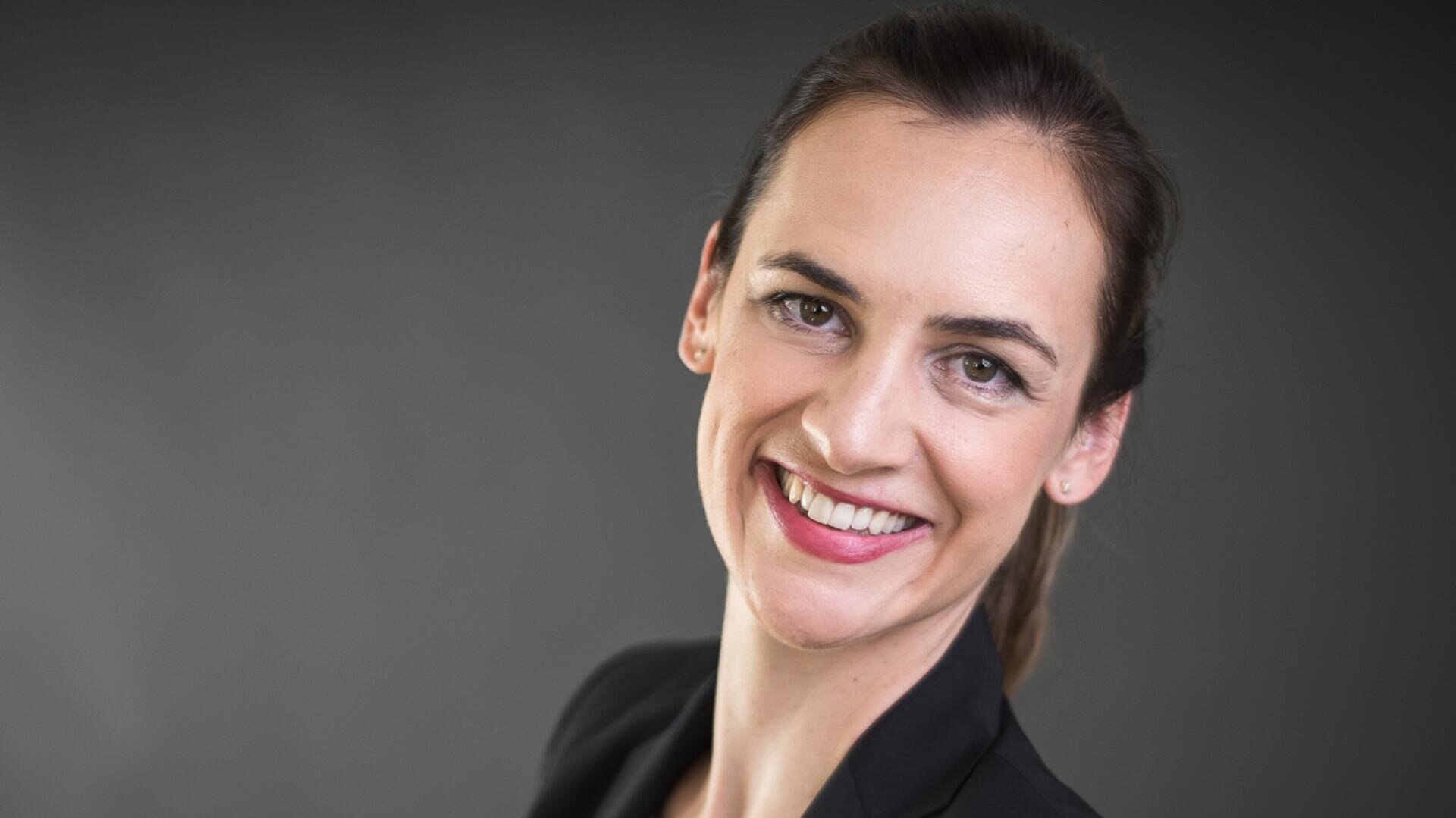 Eine Frau mit zurückgebundenem dunklem Haar lächelt in die Kamera. Sie trägt einen schwarzen Blazer und hat einen neutralen, dunklen Hintergrund hinter sich.