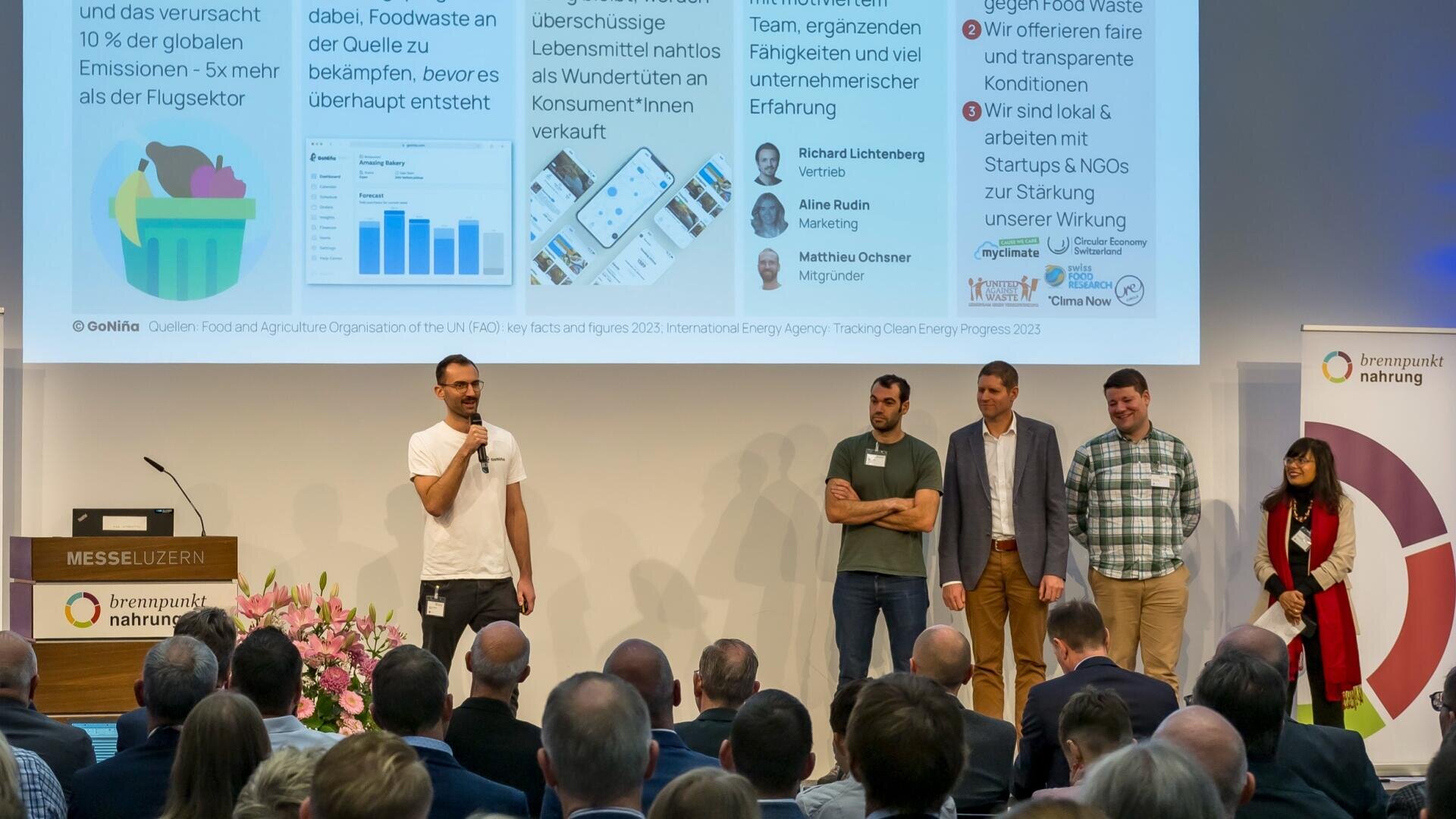 Ein Mann spricht in ein Mikrofon auf der Bühne neben vier weiteren Personen, die vor einer Präsentationsfolie stehen. Ein Publikum beobachtet sie in einem Konferenzraum mit Bannern und einem Logo mit der Aufschrift Brennpunkt Nahrung.