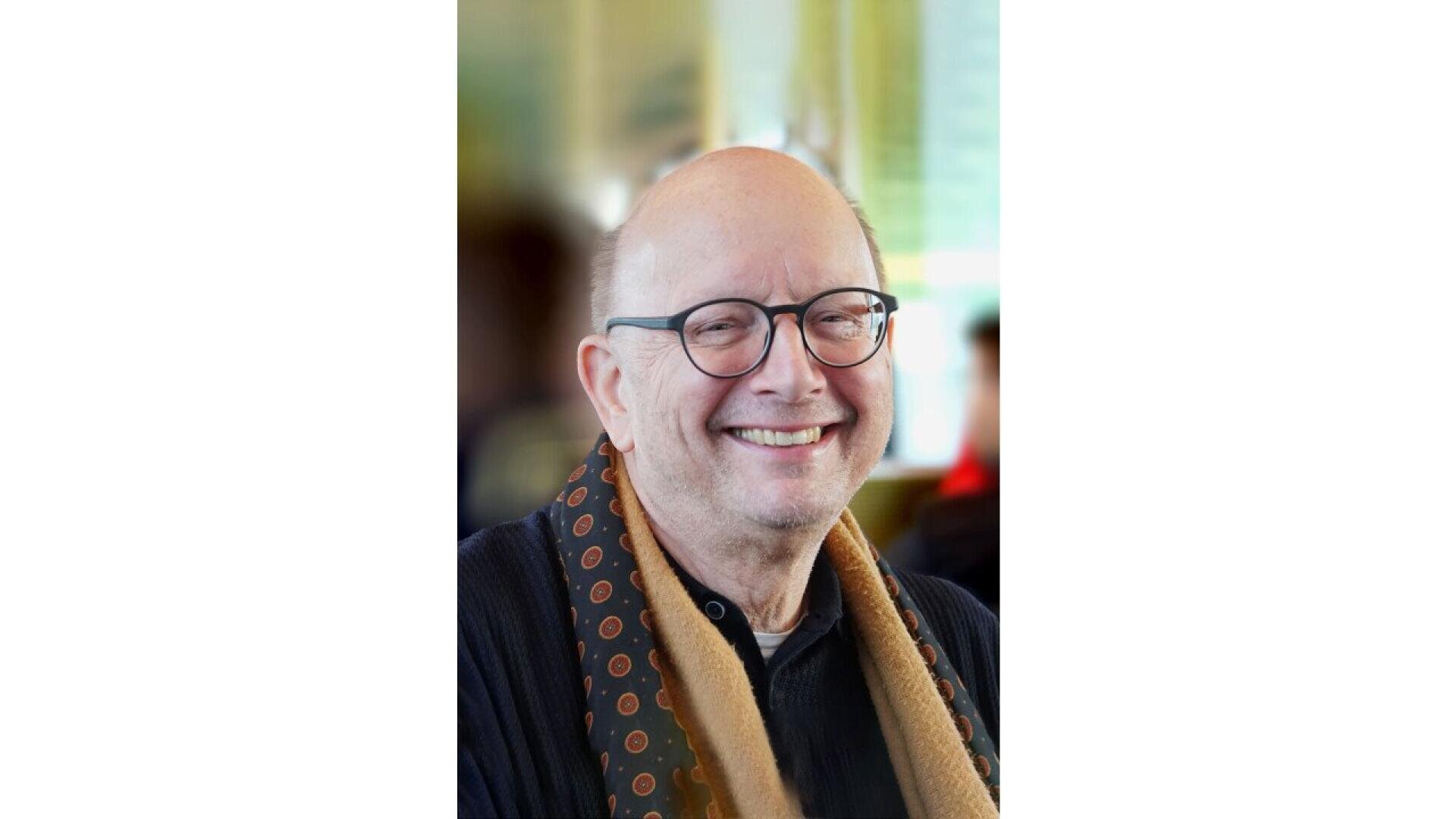 Un homme âgé souriant, portant des lunettes et une écharpe à motifs, vêtu d'un pull-over sombre, se tient à l'intérieur sur un fond légèrement flou.