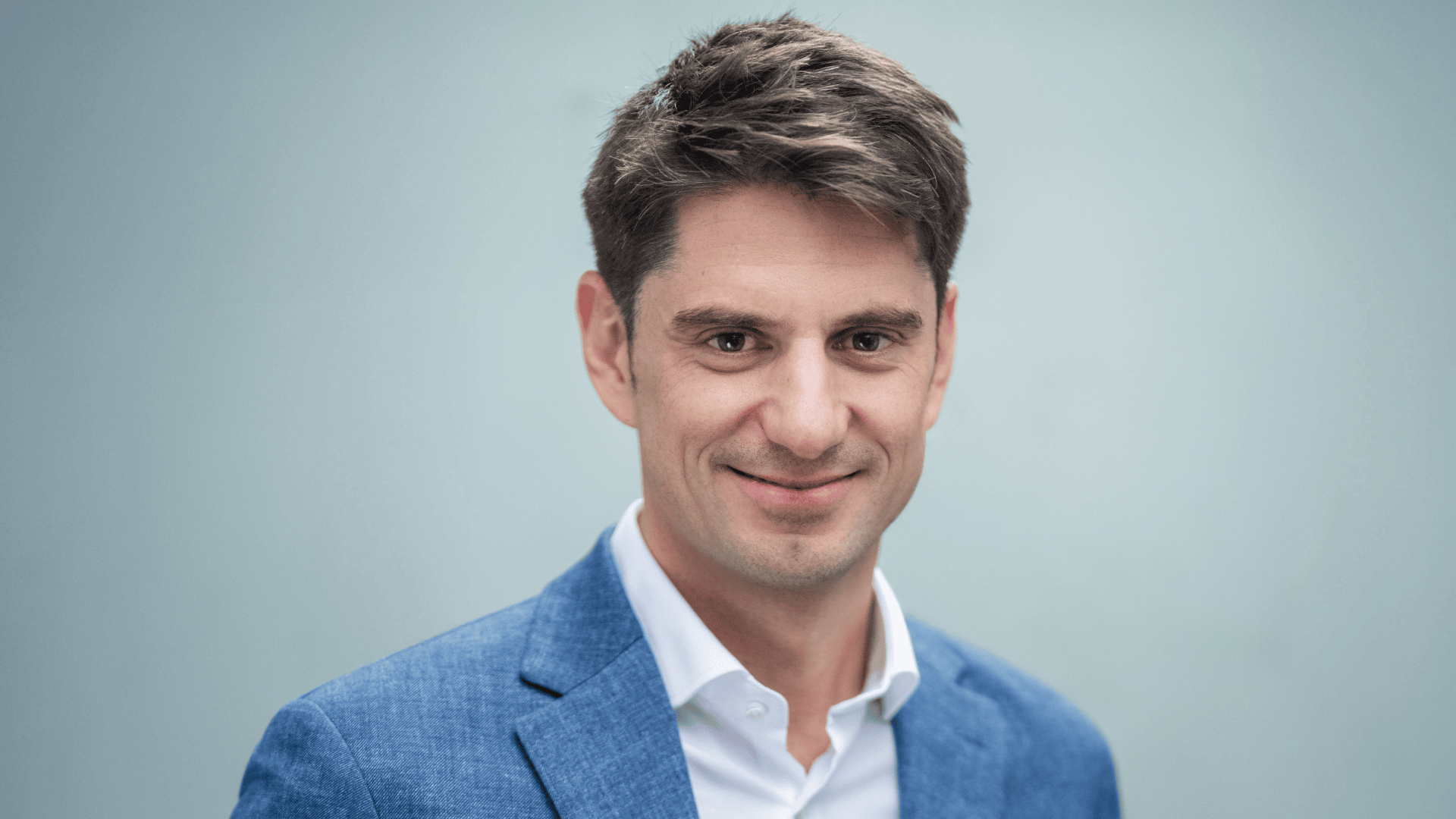 Ein Mann mit kurzen braunen Haaren, der eine blaue Anzugsjacke und ein weißes Hemd trägt, lächelt leicht, während er vor einem schlichten hellblauen Hintergrund steht.