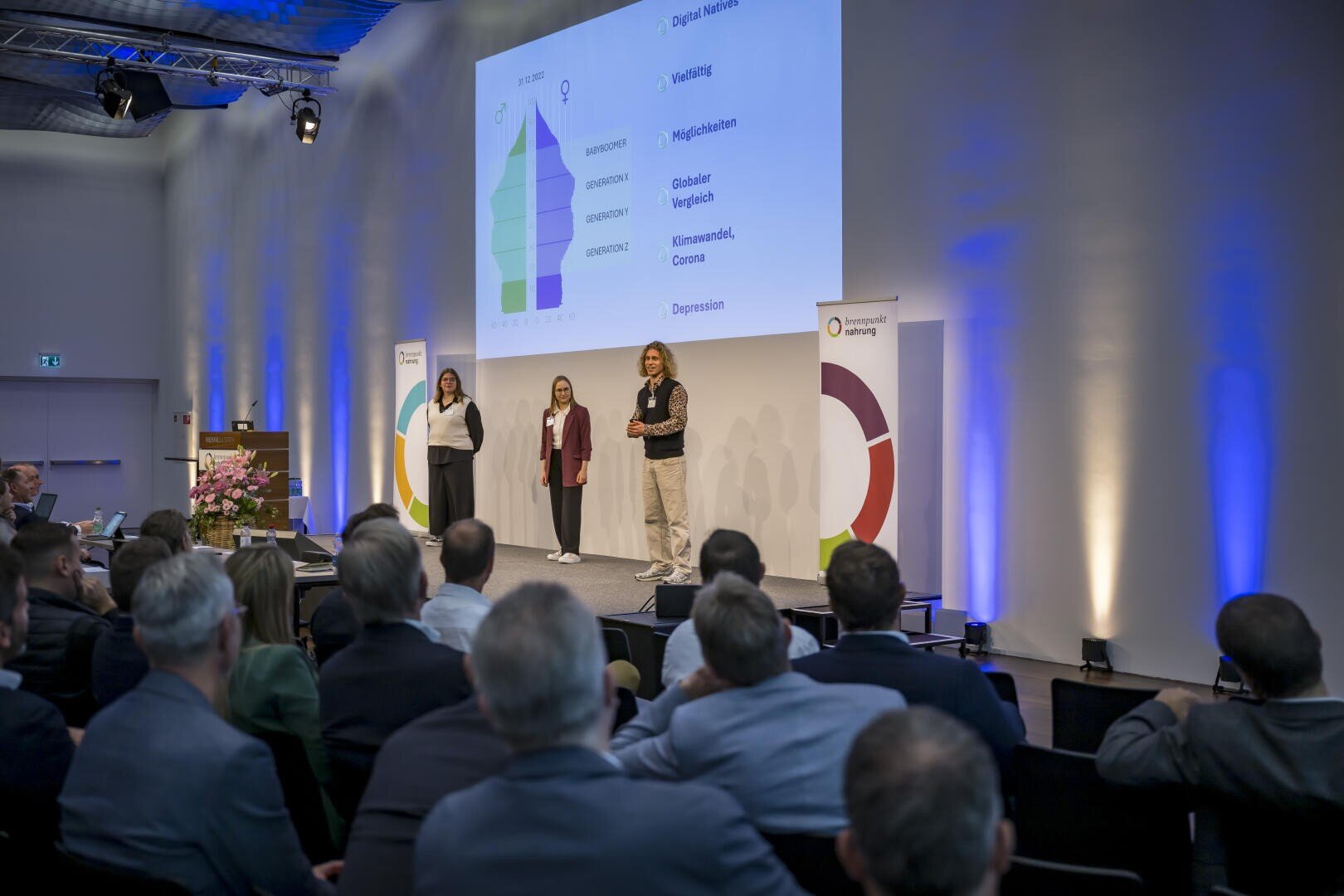 Drei Personen stehen auf einer Bühne und halten einen Vortrag vor einem sitzenden Publikum. Hinter ihnen wird eine Folie mit einem bunten Pyramidendiagramm und Text projiziert. Der Raum ist modern mit weißen Wänden und Bannern gestaltet.