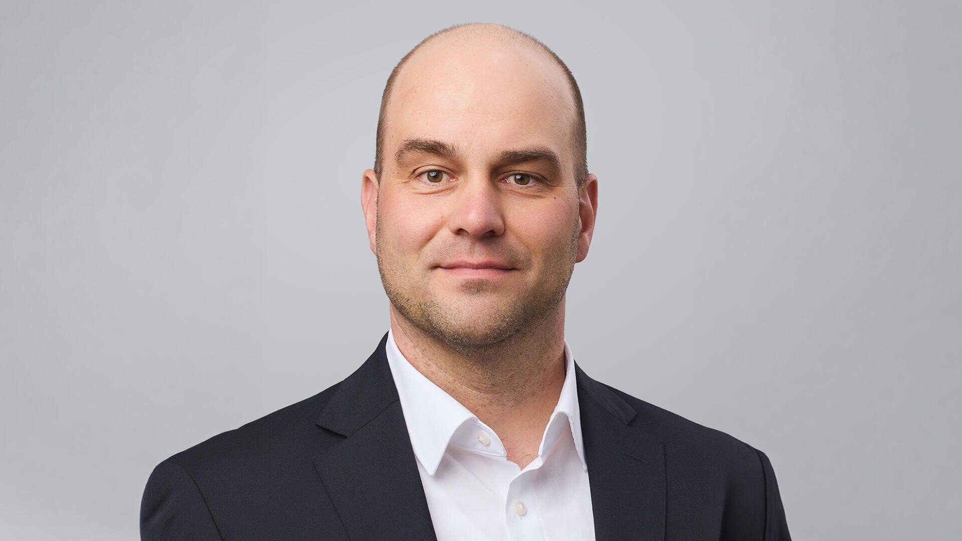 Ein Mann mit rasiertem Kopf, der ein weißes Hemd und ein dunkles Sakko trägt, steht vor einem hellgrauen Hintergrund und blickt mit einem leichten Lächeln direkt in die Kamera.