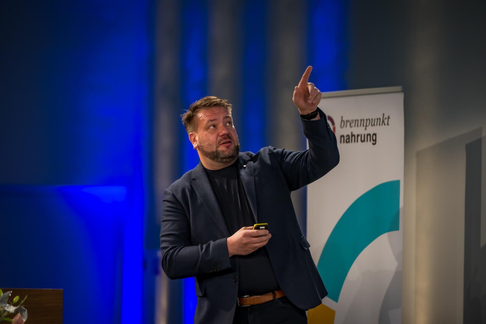 Ein Mann in einem dunklen Blazer gestikuliert mit einer Hand nach oben, während er in der anderen ein Gerät hält. Er spricht auf der Bühne neben einem Banner mit der Aufschrift brennpunkt nahrung. Blaues Licht erhellt den Hintergrund.