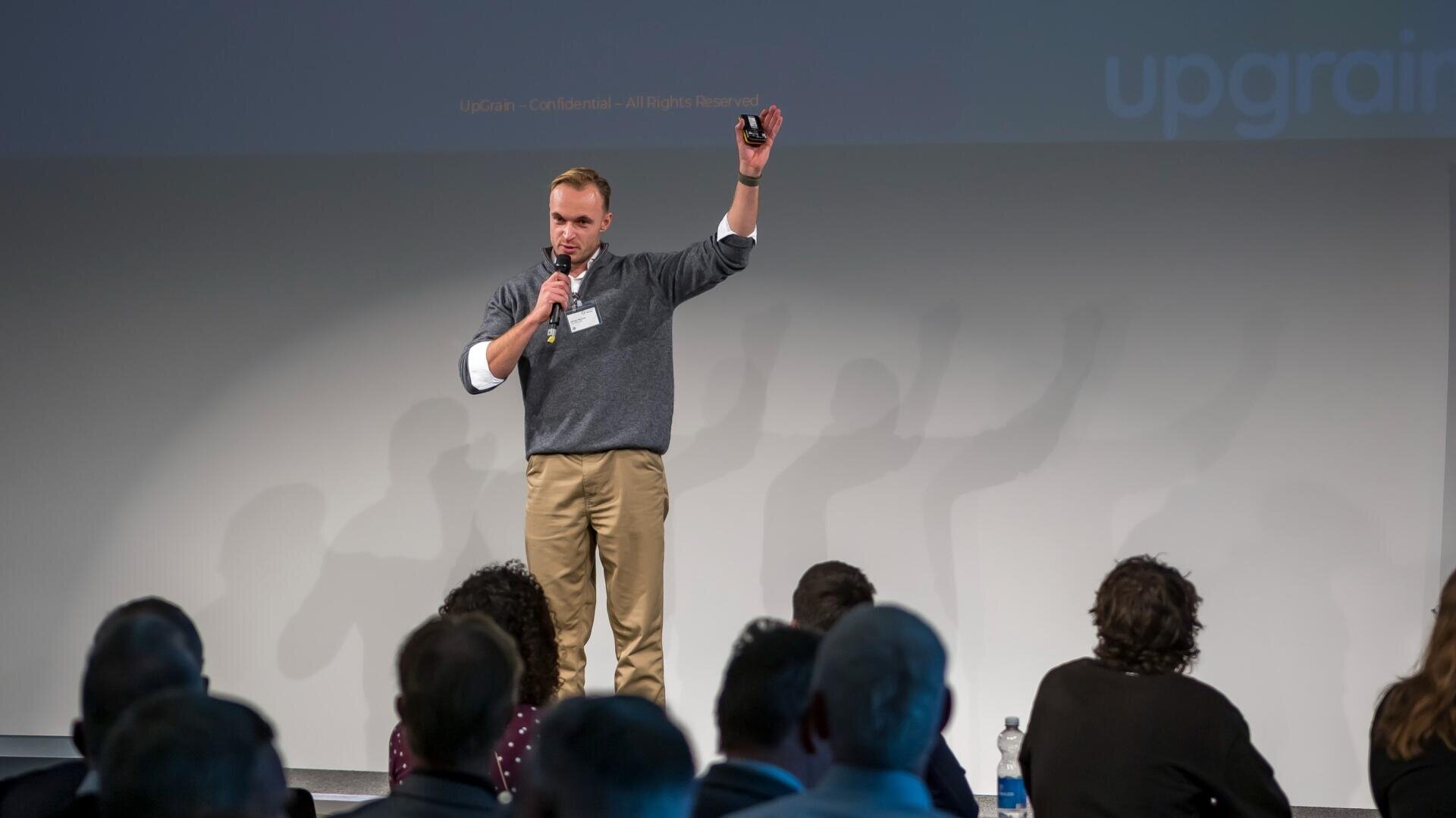 Un homme se tient sur scène, un micro à la main, et lève un bras tout en s'adressant à un public assis devant lui. Il semble donner une présentation ou une conférence dans le cadre d'un congrès.