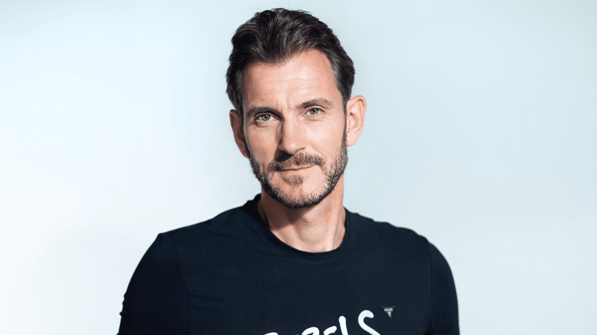 Ein Mann mit kurzen dunklen Haaren und einem gestutzten Bart trägt ein schwarzes T-Shirt und lächelt sanft. Der Hintergrund ist einfarbig und hell.