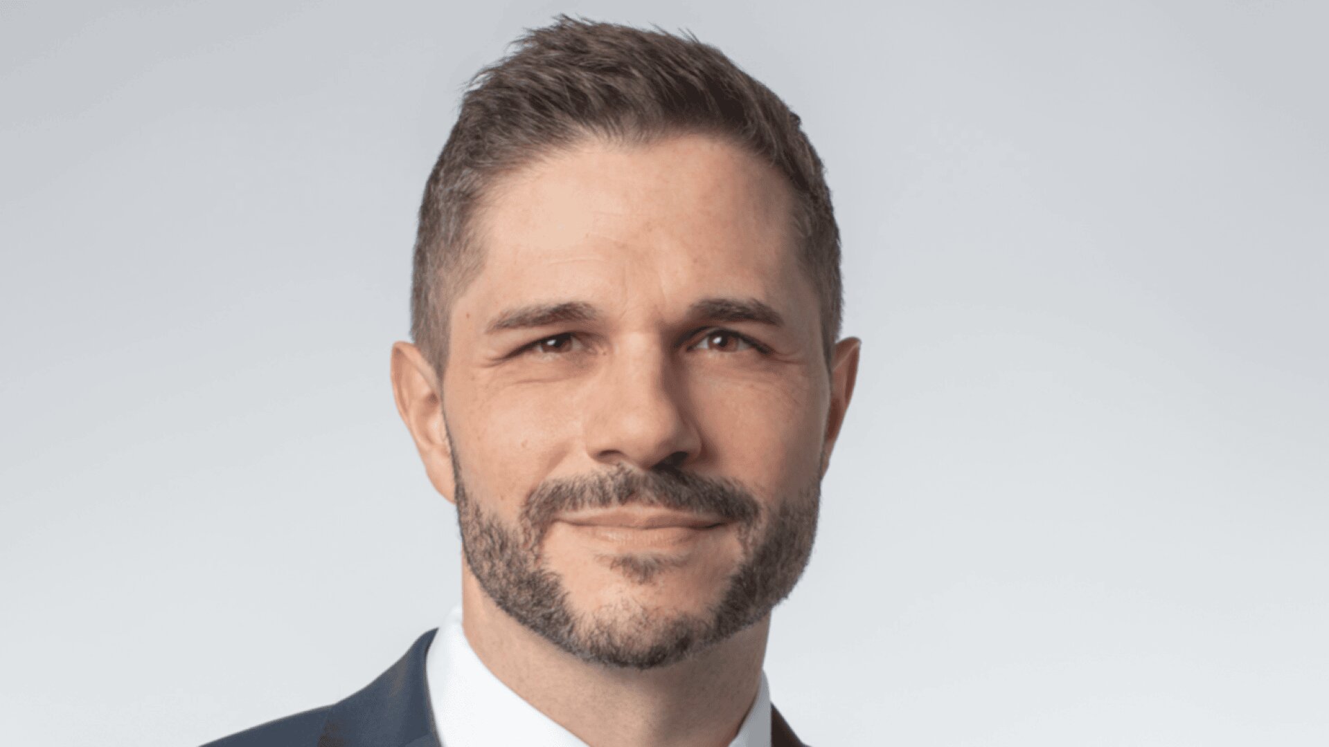 Ein Mann mit kurzen braunen Haaren und gestutztem Bart, der einen blauen Anzug, ein weißes Hemd und eine braune Krawatte trägt, lächelt leicht, während er vor einem hellgrauen Hintergrund in die Kamera blickt.