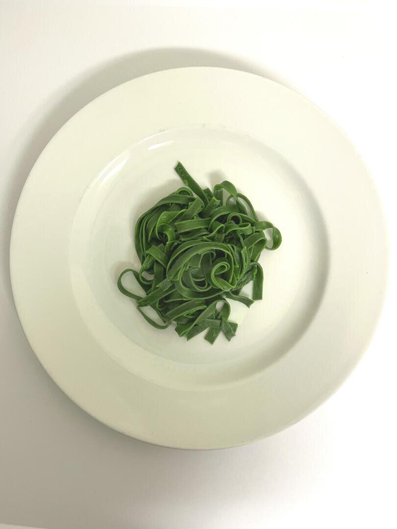 Auf einem weißen Teller befindet sich eine kleine Portion ungekochter grüner Fettuccine-Nudeln, die fein säuberlich kreisförmig auf weißem Grund angeordnet sind.