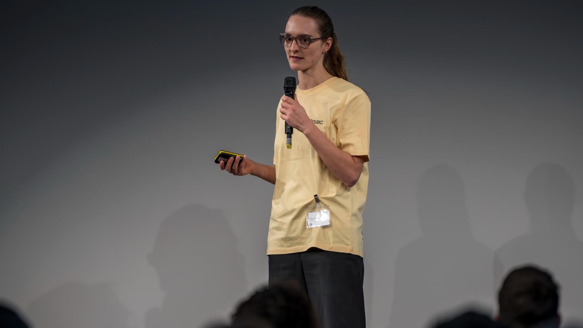 Eine Person mit langen Haaren in einem Pferdeschwanz, die eine Brille, ein gelbes Hemd und eine dunkle Hose trägt, steht auf der Bühne, hält ein Mikrofon und ein Gerät in der Hand und spricht zum Publikum.