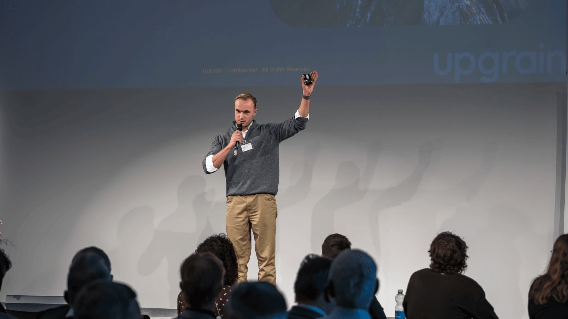 Ein Mann steht auf der Bühne, hält ein Mikrofon in der einen Hand, hebt die andere Hand und blickt in Richtung Publikum. Er scheint auf einer Veranstaltung zu präsentieren oder zu sprechen. Das Publikum sitzt und schaut aufmerksam zu.