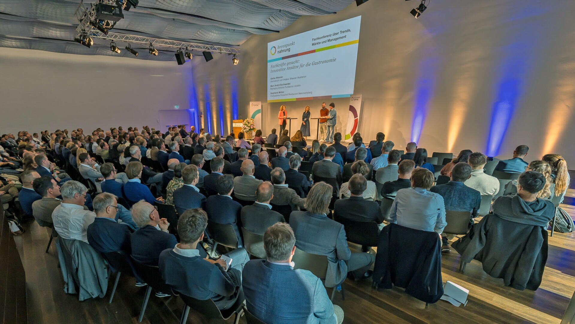 Ein großes Publikum sitzt in einem modernen Konferenzsaal und verfolgt aufmerksam eine Präsentation, die von vier Rednern auf der Bühne vor einem großen Bildschirm gehalten wird. Der Raum ist mit blauer und gelber Akzentbeleuchtung gut beleuchtet.