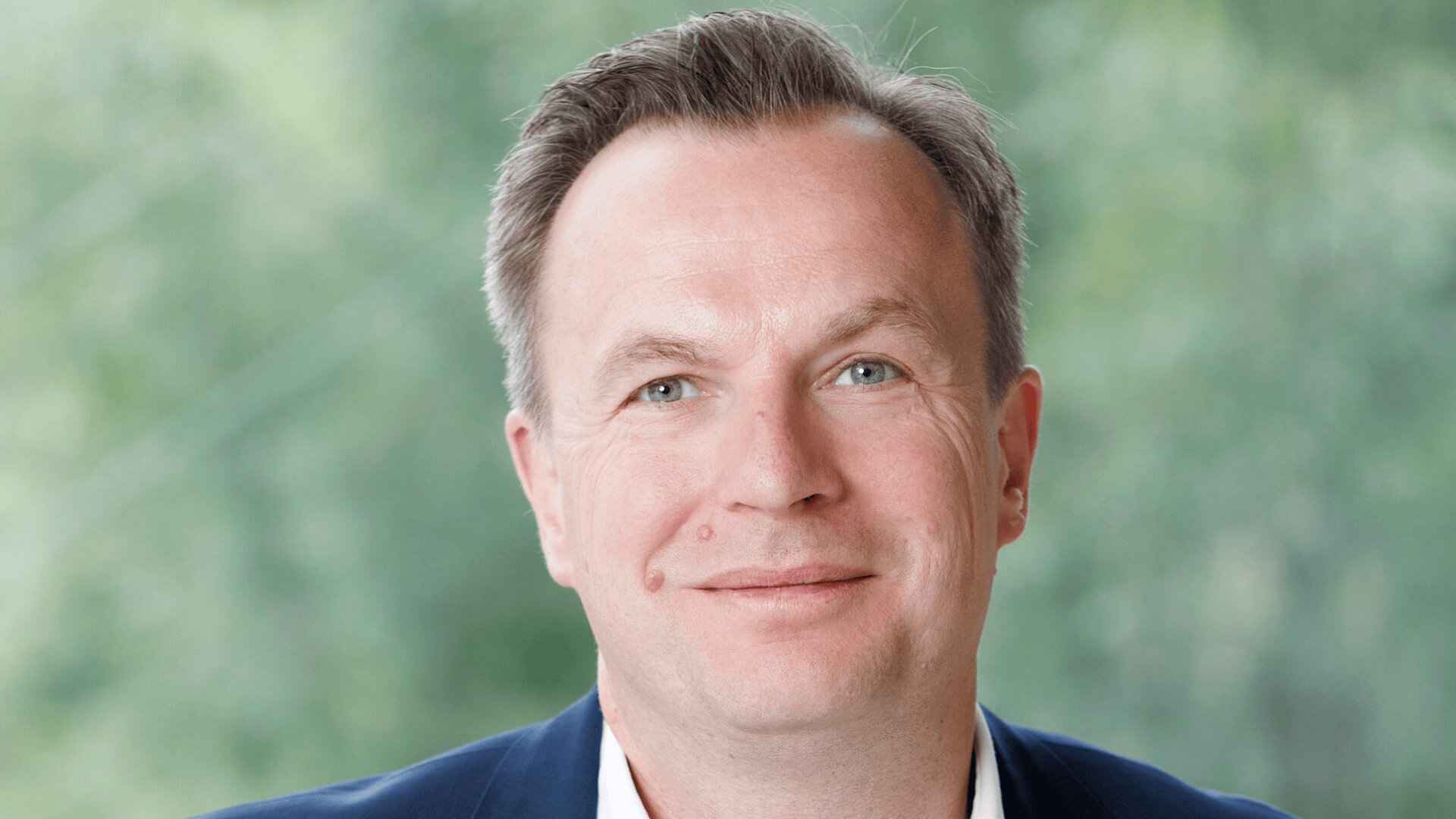 Un homme d'âge moyen aux cheveux courts châtain clair, aux yeux bleus et au léger sourire, vêtu d'un blazer foncé et d'une chemise blanche, pose sur un fond extérieur vert flou.