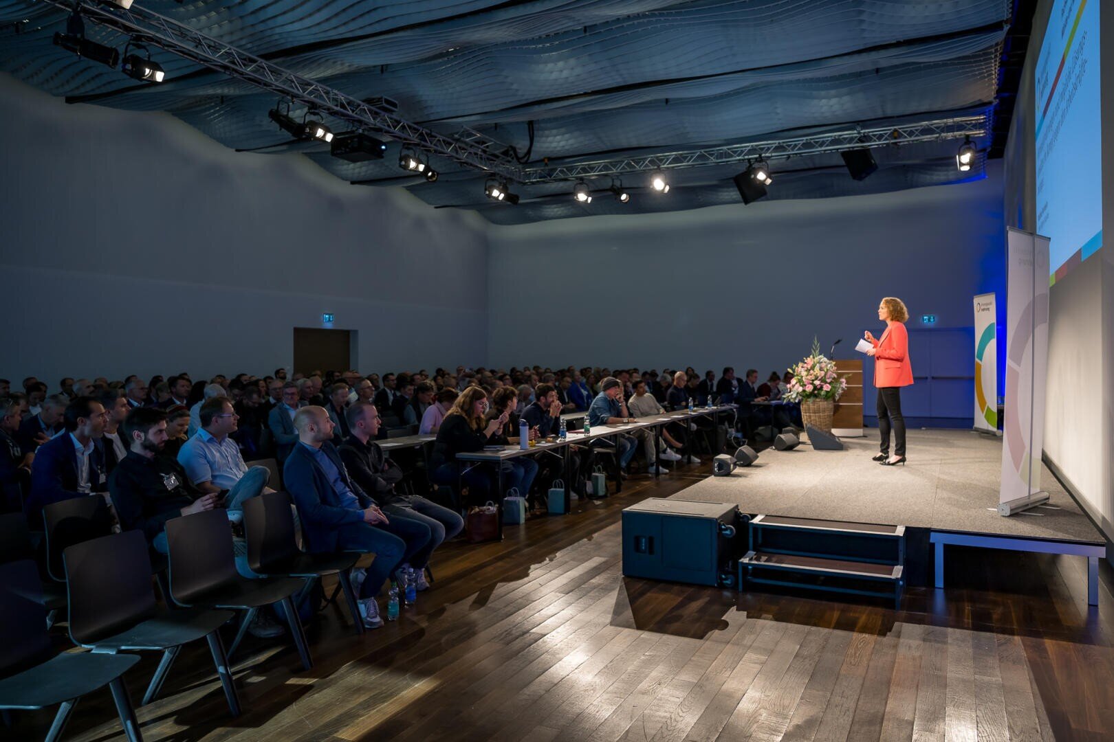 Ein Redner in einer roten Jacke steht auf der Bühne und spricht zu einem großen, sitzenden Publikum in einem modernen Konferenzraum mit hoher Decke und Bühnenbeleuchtung.