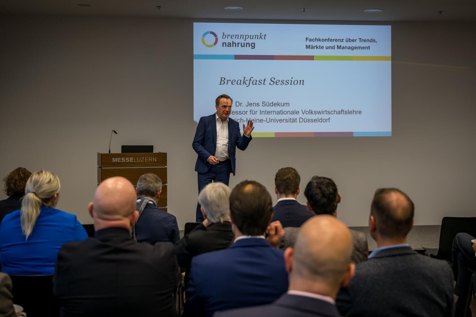 Ein Mann im Anzug spricht während einer Frühstückssitzung auf einer Konferenz zu den Zuhörern. Er steht in der Nähe eines Podiums in einem Raum mit einer Projektionsfläche, auf der sein Name und Veranstaltungsdetails angezeigt werden.
