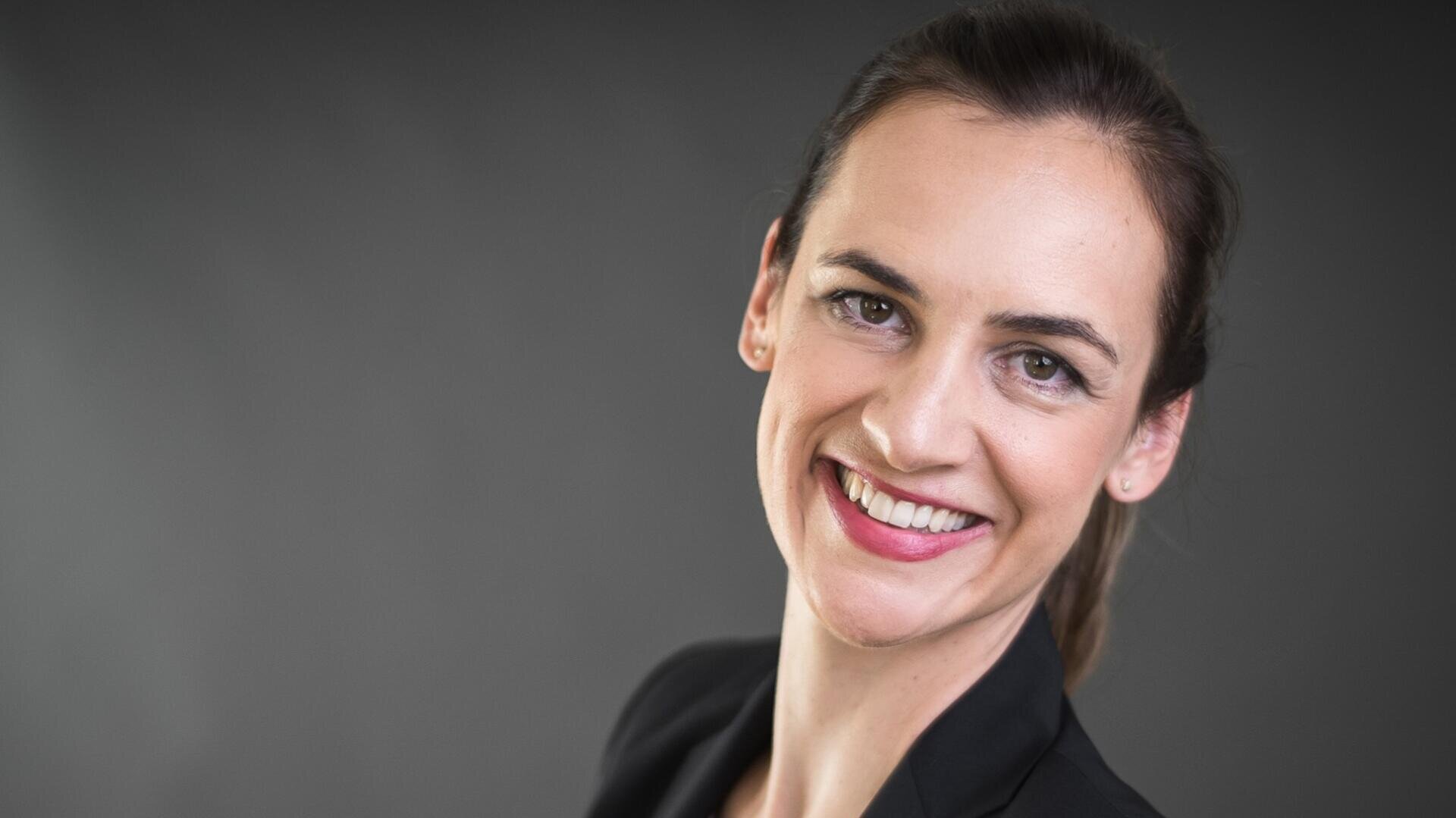Une femme aux cheveux châtains tirés en arrière sourit chaleureusement à la caméra. Elle porte un blazer noir et se tient debout devant un fond uni gris foncé.