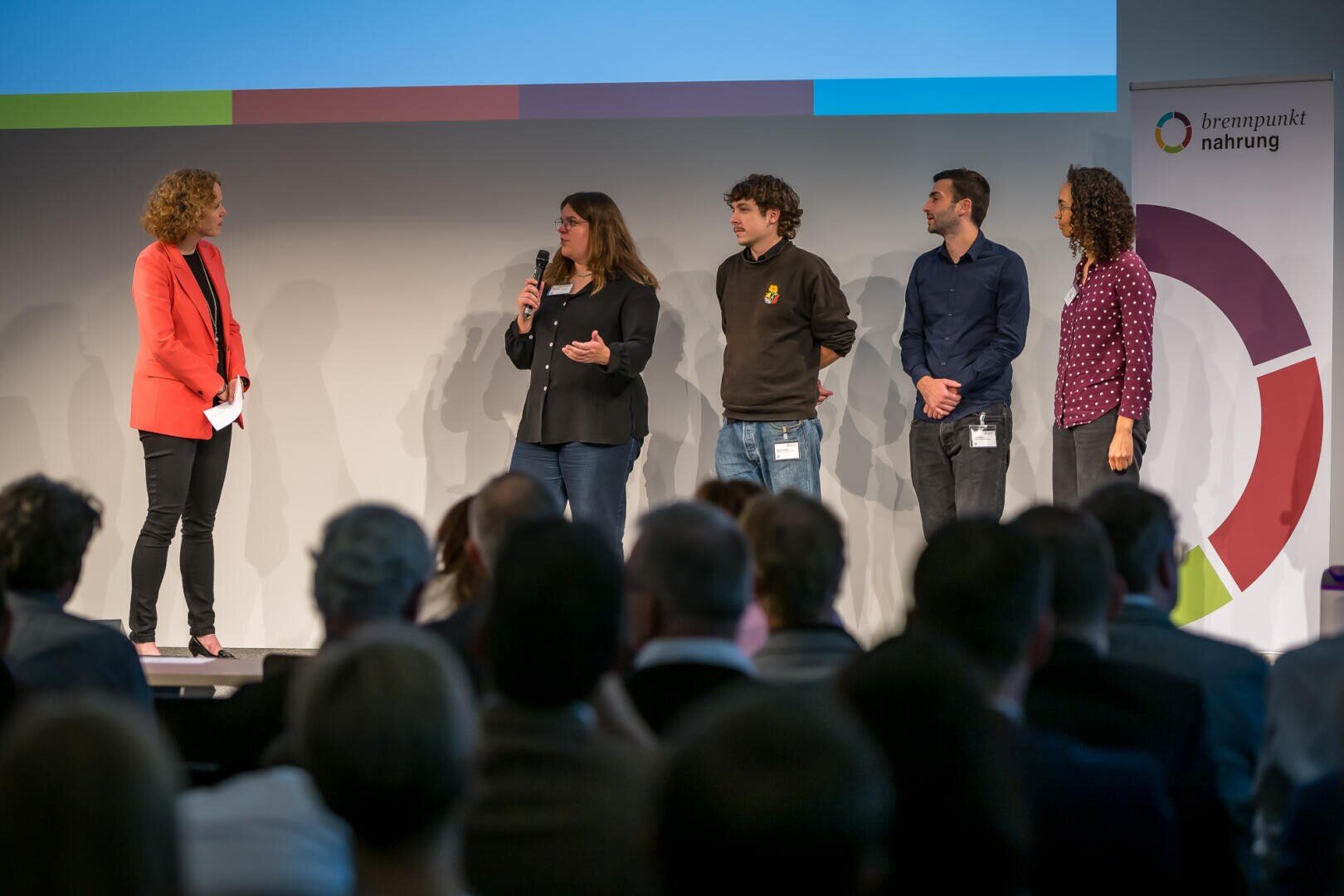 Fünf Personen stehen auf einer Bühne und führen eine Diskussion vor dem Publikum einer Konferenz. Eine Frau spricht in ein Mikrofon, während die anderen zuhören. Im Hintergrund sind ein buntes Banner und ein Logo zu sehen.