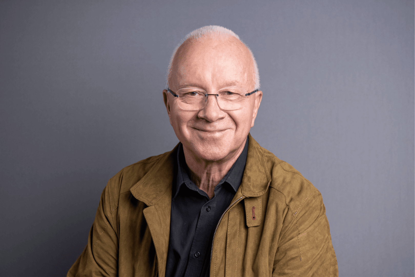 Ein älterer Mann mit kurzen weißen Haaren und Brille lächelt in die Kamera. Er trägt eine hellbraune Jacke über einem schwarzen Hemd und posiert vor einem schlichten grauen Hintergrund.