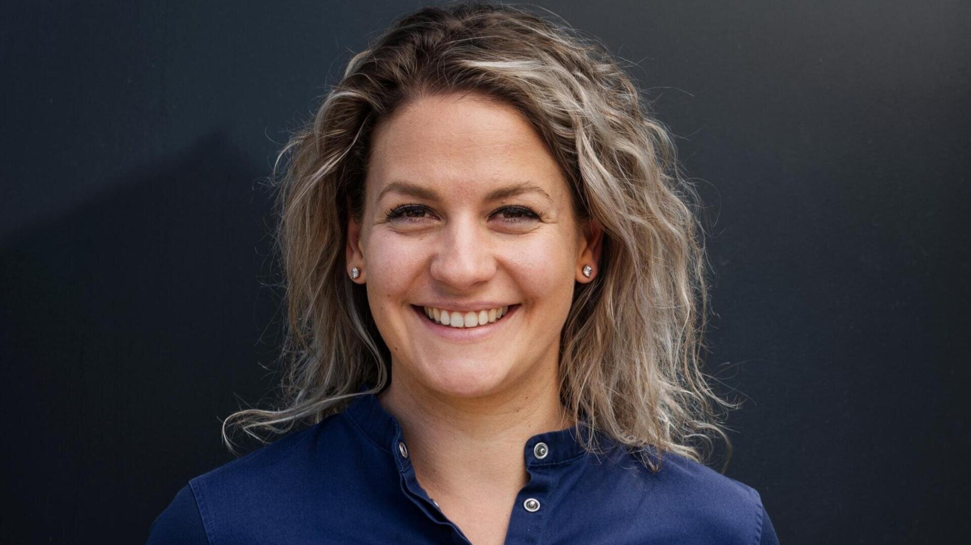 Eine Frau mit gewelltem blondem Haar und einem marineblauen Hemd mit Knöpfen lächelt in die Kamera und steht vor einem dunklen, schlichten Hintergrund.