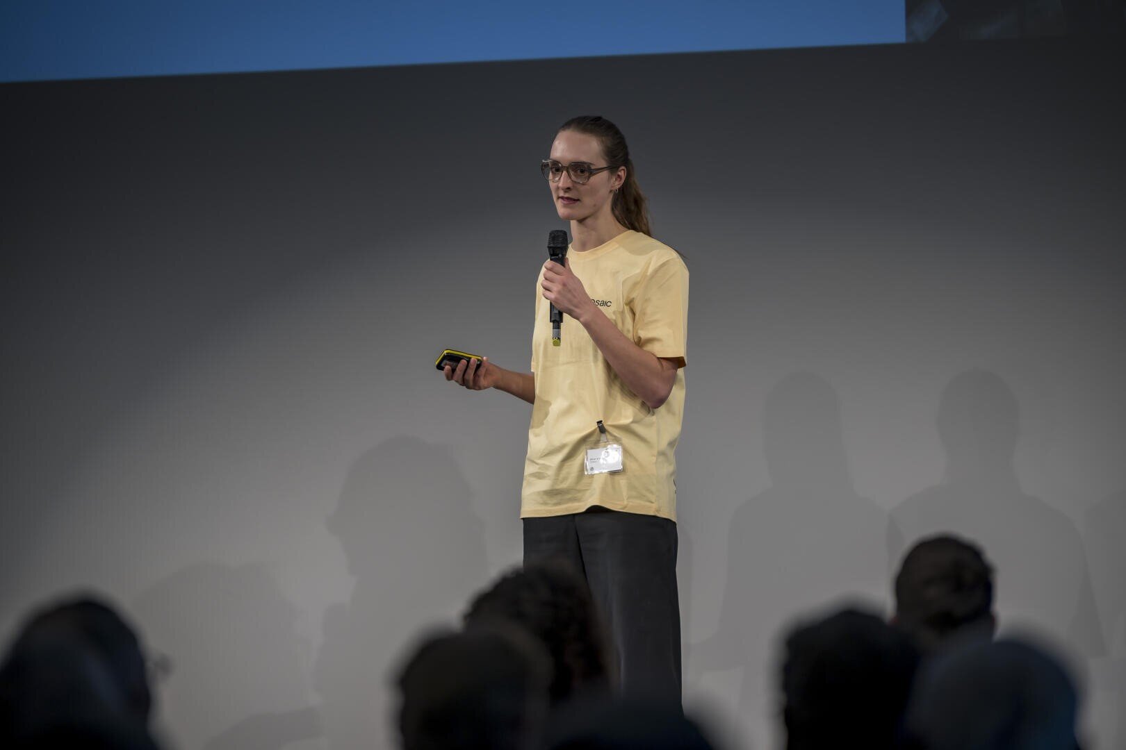 Une personne portant des lunettes et une chemise jaune se tient sur scène, un micro et un petit appareil à la main, s'adressant à un public dans une salle faiblement éclairée.