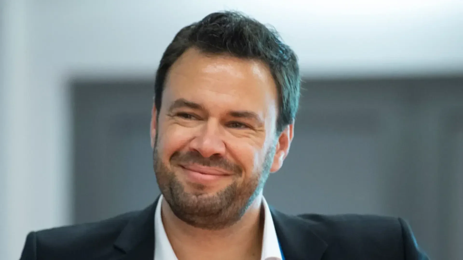 Un homme aux cheveux et à la barbe foncés, vêtu d'une veste noire et d'une chemise blanche, sourit chaleureusement. L'arrière-plan est flou et de couleur neutre.