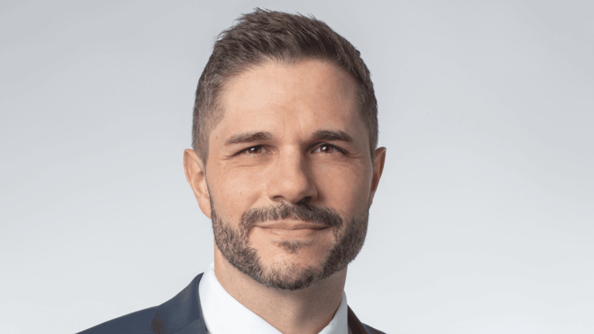 Un homme aux cheveux bruns courts et à la barbe taillée, portant un costume bleu, une chemise blanche et une cravate marron, souriant légèrement sur un fond gris clair.