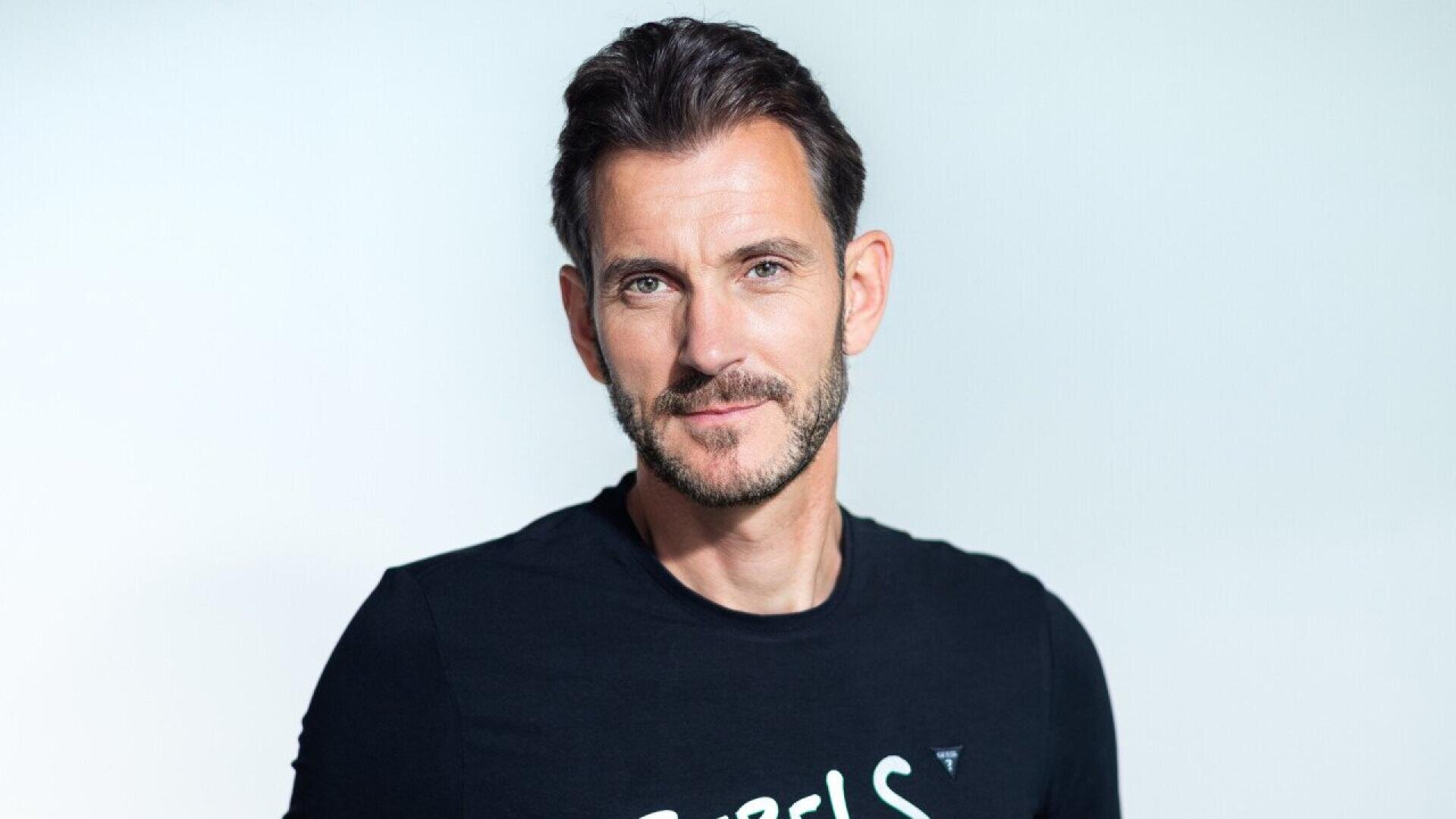 Un homme aux cheveux bruns courts et à la barbe taillée, vêtu d'un t-shirt noir, se tient sur un fond clair et uni, regardant directement l'appareil photo et souriant doucement.