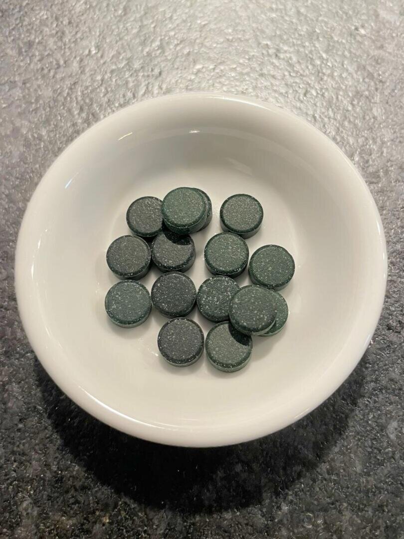 Eine weiße Schale mit dreizehn runden, dunkelgrünen Tabletten steht auf einer strukturierten, dunkelgrauen Fläche.