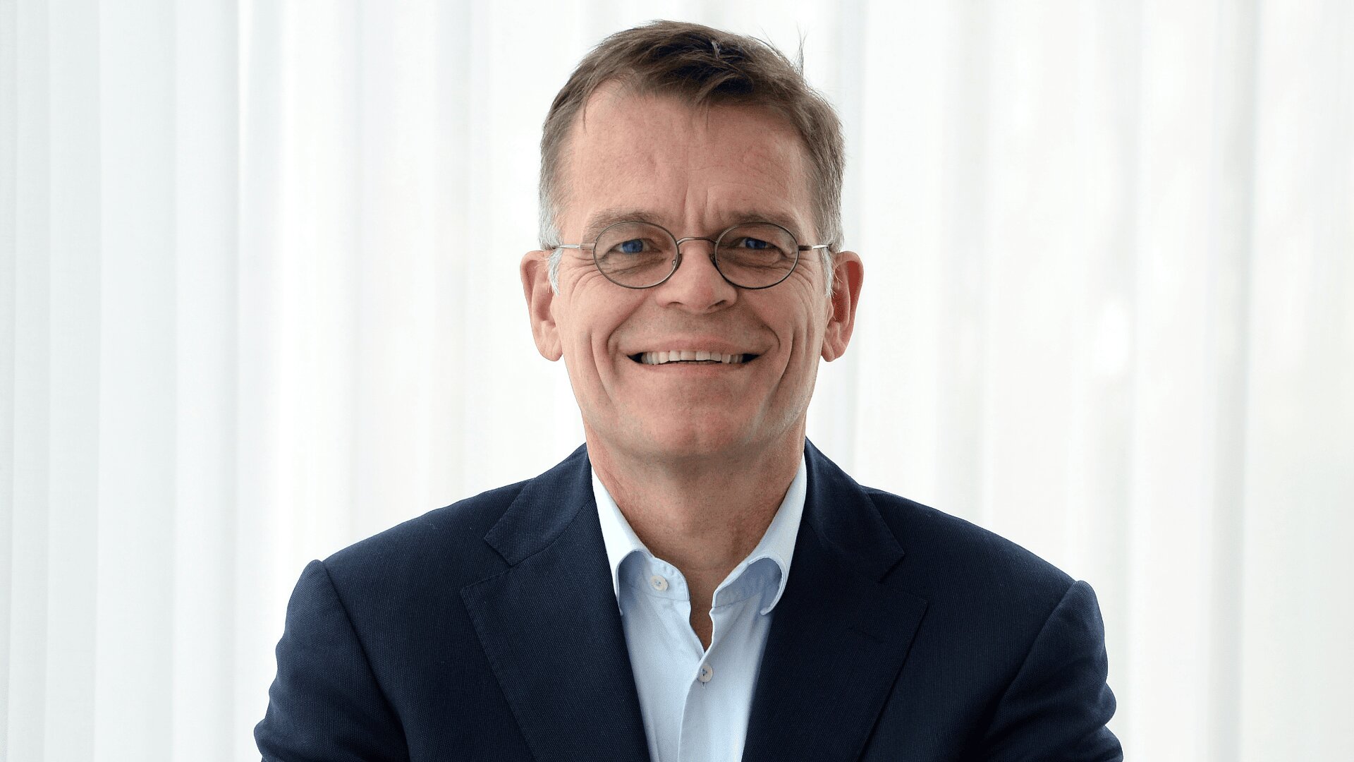 Ein Mann mittleren Alters mit kurzen braunen Haaren, der eine Brille, eine dunkle Anzugsjacke und ein hellblaues Hemd trägt, lächelt vor einem hellen, weißen Hintergrund.